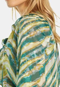Blusa trasparente con un motivo ondulato nelle tonalità di verde e giallo. Tessuto leggero con una consistenza morbida e una leggera lucentezza.