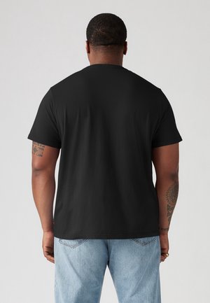 Mężczyzna ubrany w czarny krótki t-shirt i jasnoniebieskie dżinsy, stoi tyłem do kamery na jednolitym jasnym tle.