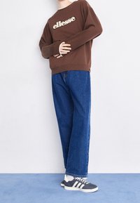 Hnědá mikina s logem "ellesse", kombinovaná s modrými širokými džíny a šedými teniskami s bílými pruhy a bílou podrážkou.