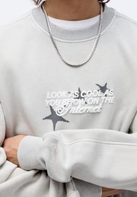 Persona che indossa una felpa grigia chiara con grafiche di stelle bianche e nere e testo "GUARDA COME SEI FICO SU INTERNET", collana d'argento visibile.