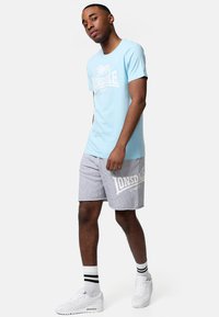 Camiseta de algodón azul claro con logo blanco, combinada con pantalones cortos grises que tienen texto blanco. Zapatos deportivos blancos y calcetines a rayas negras.