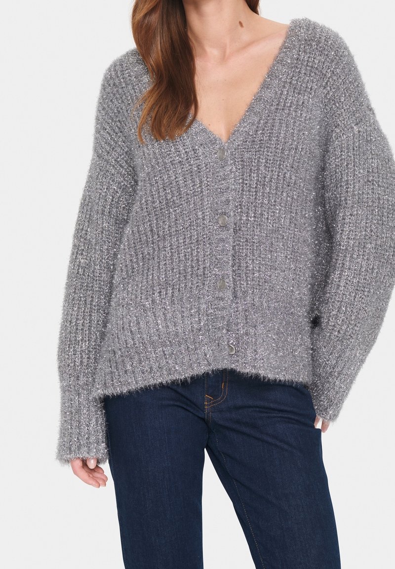 Šedý, texturovaný pletený kardigan s výstřihem do V, oversized střihem a velkými knoflíky. Má volné pletení a mírný lesk, naložený na tmavé džíny.