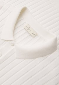 Polo blanc en maille côtelée avec col plié, patte de boutonnage et étiquette visible de taille et de marque à l'intérieur du col.