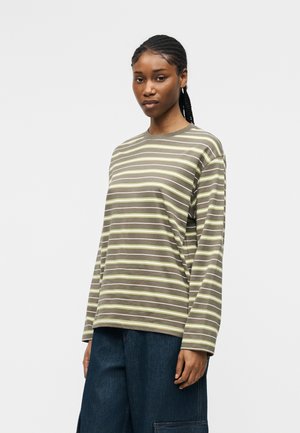 Žena s vrkočmi, oblečená do olivovozelenej a žltej pruhovanej dlhorukávovej košele a tmavomodrých širokých nohavíc, stojaca pred jednofarebným pozadím.