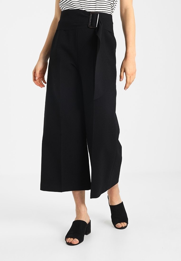 Femme portant un pantalon noir large taille haute raccourci avec une ceinture en tissu et des mules noires à talons blocs ouverts à l'avant, debout sur un sol blanc.