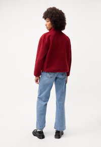 Rød fleecejakke med høy krage, avslappet passform og teksturert overflate; kombinert med lyseblå jeans og svarte kraftige sko.