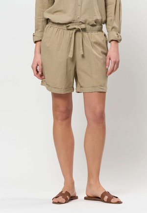 Kvinne iført beige linshorts med snor i midjen og brune flate sandaler, stående foran en ensfarget bakgrunn.