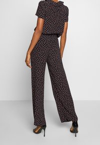 Combinaison noire avec motif de cœurs roses, manches courtes, coupe décontractée, taille haute et pantalon à jambes larges. Portée avec des sandales à talons hauts noires.