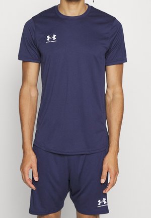 Mężczyzna ubrany w granatowy sportowy t-shirt i szorty marki Under Armour, stojący na tle jednolitego, jasnego tła.