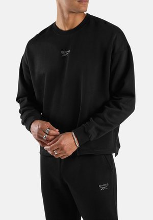 Mann trägt schwarzen Reebok-Sweatshirt und passende Sweatpants, Hände mit mehreren Ringen und Armband verschränkt, blickt zur Seite.