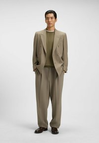 Beige doppelreihiger Anzugjacke mit zwei-Knopf-Verschluss, passenden weit geschnittenen Hosen, kombiniert mit einem olivgrünen Pullover und braunen Lederschuhen.