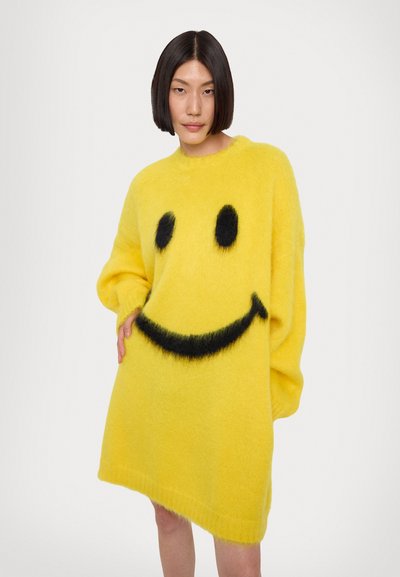 Abito maglione giallo fluffy con un grande design di faccina sorridente nera, con maniche lunghe e vestibilità ampia. Texture morbida e scollatura rotonda.