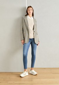 Rutig kavaj i beiget och svart med två fickor, lager på lager över en ljusbeige stickad tröja, matchad med blå skinny jeans och vita sneakers.