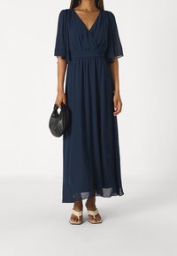 Marinblå maxiklänning med V-ringning, utställda ärmar och cinchad midja. Bärs med krämfärgade sandaler och en svart kuvertväska.