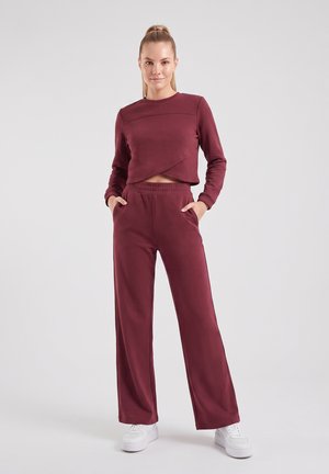 Donna in piedi con le mani nelle tasche, indossa un crop top bordeaux a maniche lunghe e pantaloni a gamba larga abbinati con sneakers bianche su sfondo semplice.