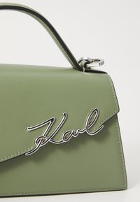 Πράσινη τσάντα χερούλι από τεχνητό δέρμα με δομημένο σχέδιο, μεταλλικό λογότυπο και αντίθετη μαύρη περίμετρο. Διαθέτει χερούλι στην κορυφή και γωνιώδη κλείσιμο με καπάκι.