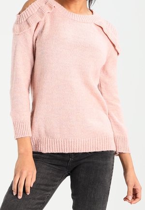 Femme portant un pull rose clair à épaules dénudées avec poignets et ourlet côtelés, associé à un jean gris foncé sur un fond uni.