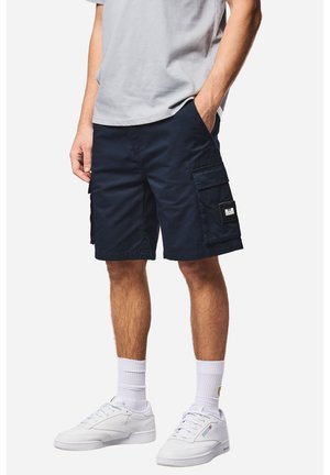 Hombre vistiendo pantalones cortos cargo azul marino, camiseta gris claro, calcetines blancos y zapatillas blancas, de pie con una mano en el bolsillo.