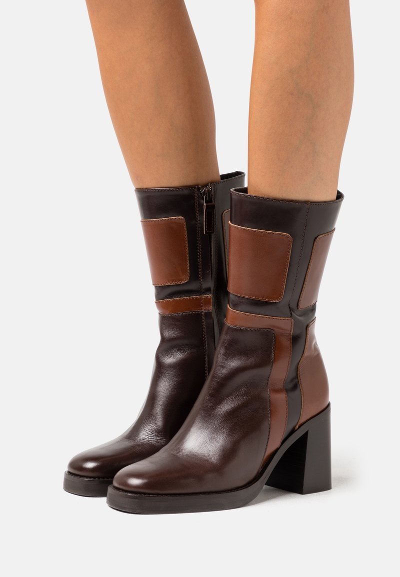 Bottes en cuir marron foncé mi-mollet avec talons blocs, patches rectangulaires marron clair et fermetures éclair latérales, portées sur jambes nues.