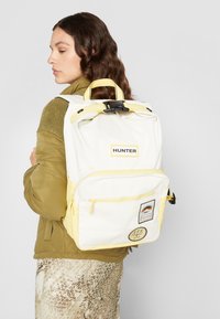 En kvinna med långt, vågigt hår bär en grön jacka och en vit och gul ryggsäck med en logotyp, ser åt sidan mot en neutral bakgrund.