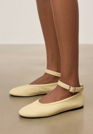 Pieds portant des ballerines en cuir jaune pâle avec des brides autour de la cheville et des boucles argentées sur un sol beige lisse.