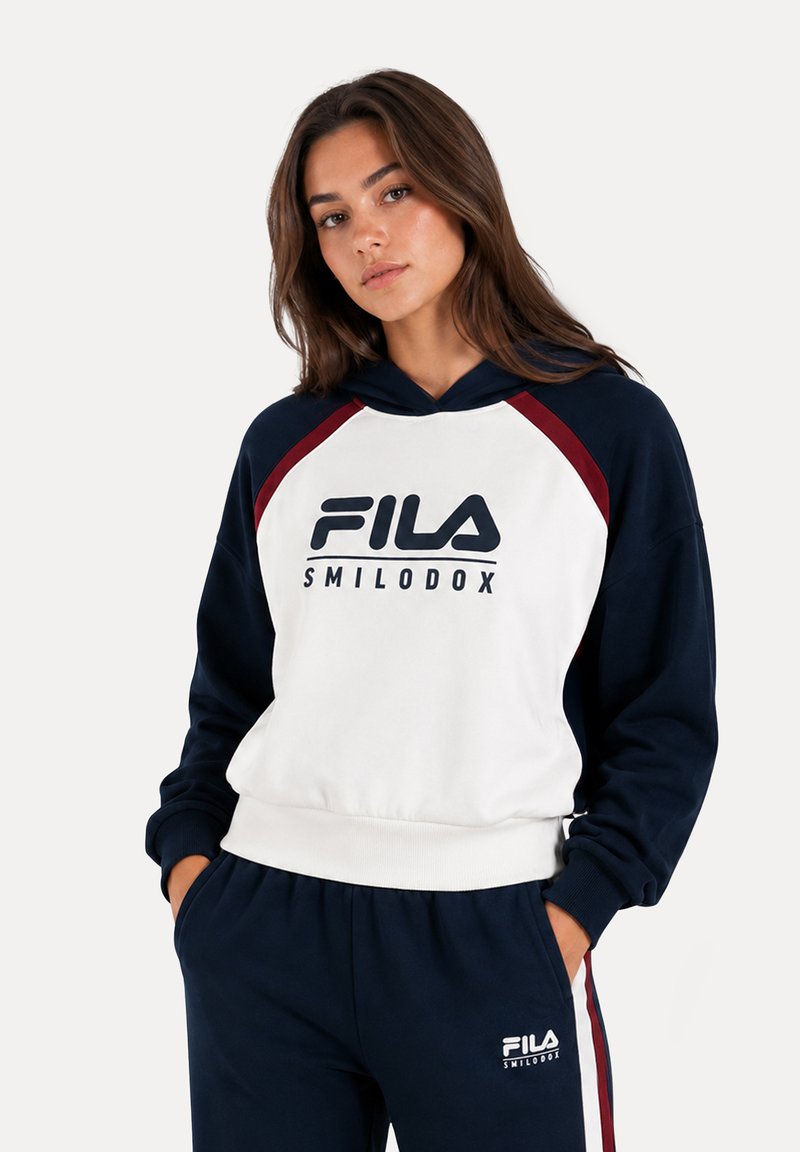 Femme portant un sweat à capuche FILA Smilodox blanc et marine et un pantalon de survêtement marine, debout, les mains dans les poches, regardant légèrement sur le côté.