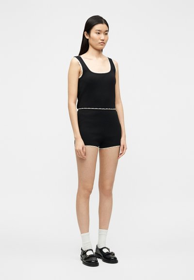 Jeune femme debout portant un haut noir sans manches et un short assorti avec un liseré blanc, des chaussettes blanches et des chaussures noires à boucle sur un fond uni.