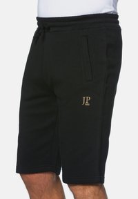Pantalones cortos de sudor negros fabricados con material suave, con una cinturilla elástica con cordón y bolsillos laterales, adornados con un detalle de logo dorado.