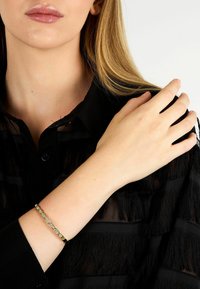 Bracciale d'oro con maglie testurizzate, posato su una blusa nera con frange. Presenta una chiusura a moschettone, che ne evidenzia il design delicato.