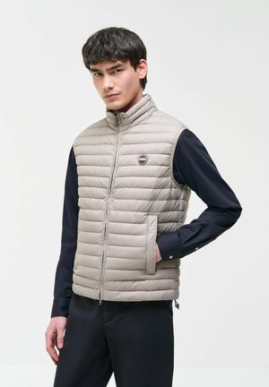Giovane uomo che indossa un gilet trapuntato beige senza maniche sopra una maglia nera a maniche lunghe, in piedi con una mano nella tasca del gilet, espressione neutra.