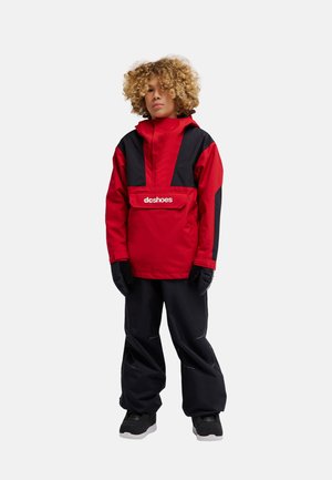 Kind met krullend haar dat een rood-zwarte jas draagt, zwarte handschoenen, zwarte broek en zwarte schoenen, staand tegen een effen achtergrond.