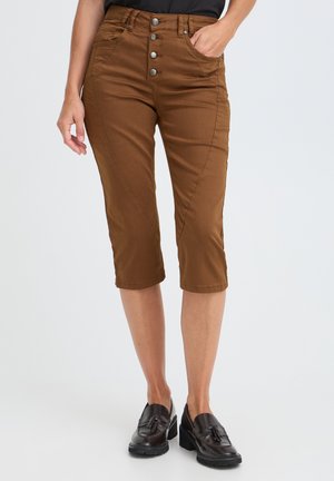 Femme portant un pantalon court marron taille haute avec boutons sur le devant et des mocassins noirs, sur un fond clair uni.