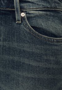 Detailní záběr na kapsu modrých džínsů, zobrazuje poutko na opasek, nýtek, šití a texturu látky.