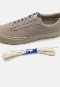 Sneaker in suede beige con finitura liscia, punta rotonda e suola in gomma spessa. Include lacci piatti bianchi avvolti in una combinazione di blu e bianco.