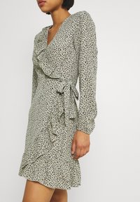 Robe verte à encolure en V, manches longues, accents froncés et motif éclaboussé de petits points noirs. Le tissu semble léger et texturé.