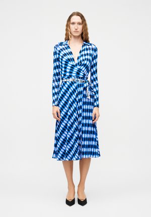 Mujer de pie con un vestido cruzado azul con estampado, mangas largas, cinturón de lazo y tacones negros de punta, sobre un fondo blanco liso.