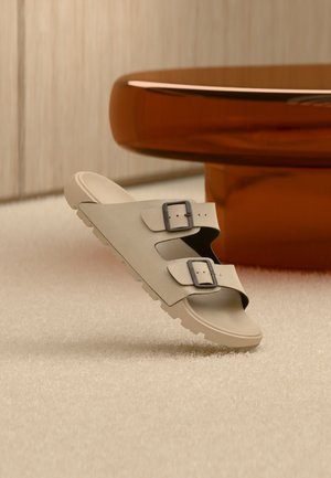 Sandale beige avec deux boucles noires en équilibre sur un sol blanc texturé, devant une table ronde et brillante couleur ambre, dans une pièce minimaliste.
