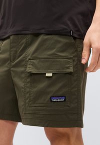 Κοντινή λήψη χακί cargo σορτς με τσέπη με καπάκι και λογότυπο Patagonia που φοριέται από άτομο που στέκεται με μαύρο πουκάμισο.