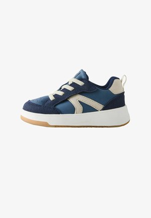 Low-top sneaker with navy and beige suede and fabric upper, white laces, thick white sole, and light brown rubber outsole.