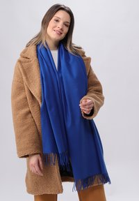 Écharpe bleue en tissu doux, ornée de franges. Portée avec un manteau teddy texturé beige. Design simple et rectangulaire.