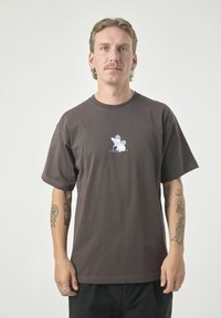 Brown cotton t-shirt featuring a small light purple cartoon graphic on the chest. It has short sleeves and a relaxed fit.