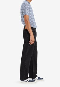 Adulte portant une chemise à manches courtes rayée bleu et blanc, un jean noir droit et des baskets bleu et blanc, debout de côté.