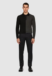 Uomo che indossa un polo a maniche lunghe bicolore nero e grigio scuro, pantaloni grigio scuro e scarpe di pelle nere, in piedi davanti a uno sfondo neutro.
