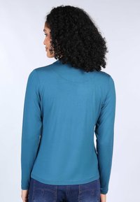 Long-sleeve top in teal, made of smooth fabric. Features a mock neck and subtle curved seam detailing at the back. Paired with denim.