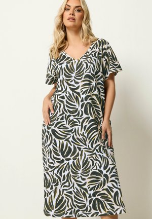 Kvindte iført en løs maxi-kjole med korte ærmer med grønne og sorte tropiske bladprint og V-udskæring, stående mod en ensfarvet lys baggrund.