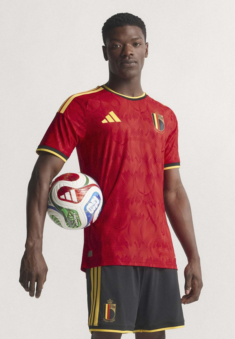 Crvena nogometna majica s teksturiranim dizajnom, crne kratke hlače s žutim prugama i nogometna lopta u više boja držana pod jednim okom.