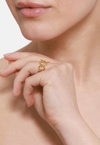 Anello d'oro con elementi circolari collegati. Texture liscia, design aperto. Indossato su una mano con una tonalità della pelle chiara su uno sfondo neutro.