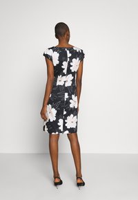 Mujer de pie de espaldas, vestida con un vestido negro de longitud hasta la rodilla con grandes estampados florales en blanco y tacones negros tipo slingback.