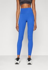 Høytlivede blå leggings laget av elastisk materiale, med en jevn tekstur og en tettsittende design. Båret med svarte treningssko.