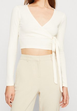 Vrouw draagt een wit, cropped wikkelshirt met lange mouwen dat aan de zijkant is gestrikt, gecombineerd met een beige broek met hoge taille.